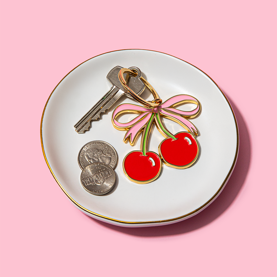 Keychain with cherries and a bow on a white dish with keys and coins against a pink background