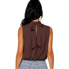 A woman shown from the back wearing a sleeveless brown top with a large bow detail at the back neckline, paired with light blue jeans against a plain white background.