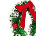 A close-up of a green tinsel Christmas wreath decorated with a large red velvet bow, a red ornament, and silver bells on a white background.