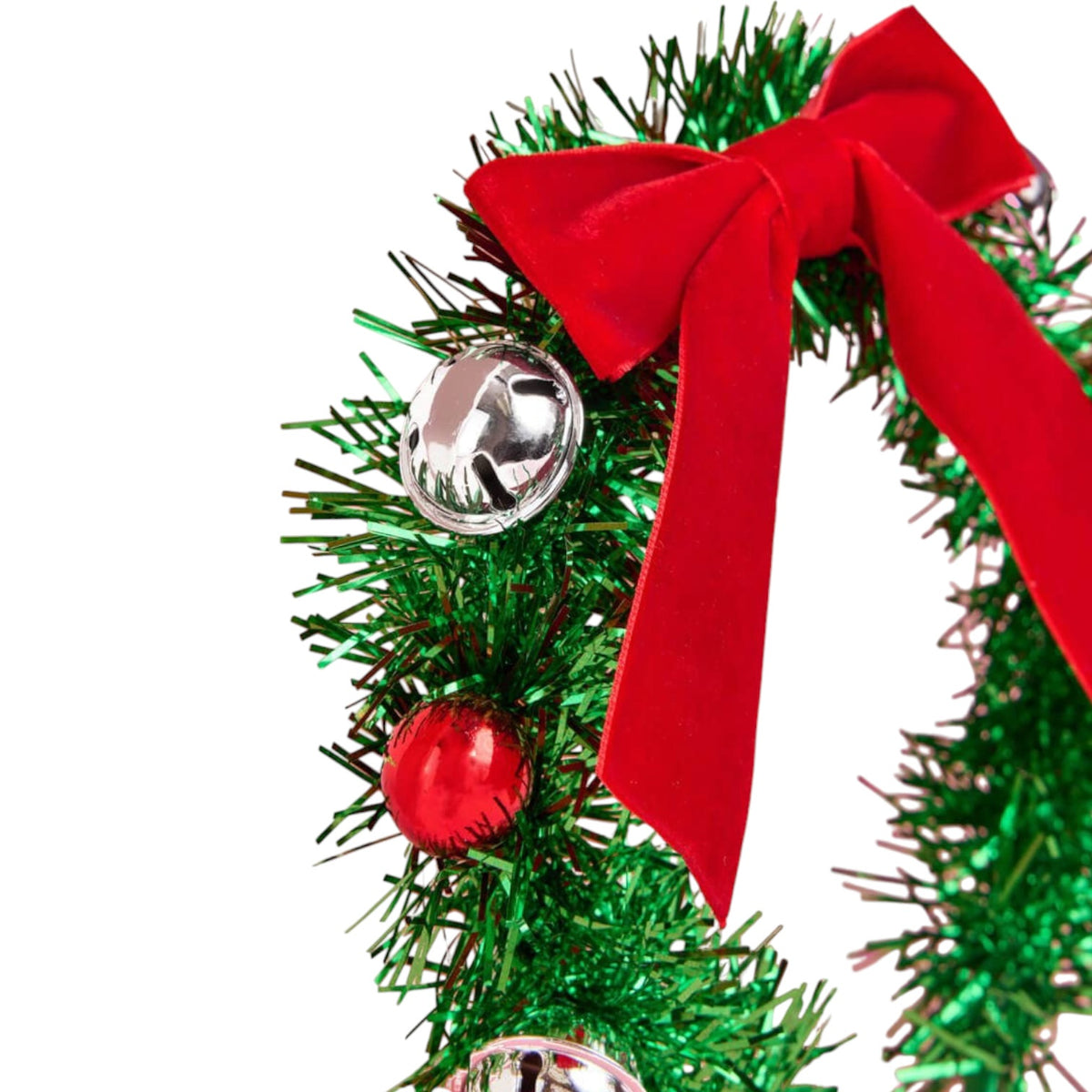 A close-up of a green tinsel Christmas wreath decorated with a large red velvet bow, a red ornament, and silver bells on a white background.
