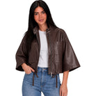 A woman modeling a cropped brown faux-leather jacket with wide sleeves, a front zipper, and large chest pockets, worn over a white top and blue jeans against a plain white background.