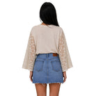 A woman shown from the back wearing a beige long-sleeve top with sheer lace sleeves and a blue denim mini skirt, against a plain white background.