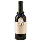 A dark glass wine bottle with a blank cream label, decorated with a blue beaded necklace and an oval anchor-patterned pendant, on a plain white background.