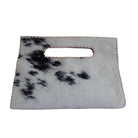 A rectangular handbag made from cowhide with a black and white spotted pattern, featuring a cutout handle and light-colored contrast stitching, shown against a plain white background.