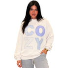 A woman modeling a cream-colored crewneck sweatshirt with 'COZY' written across the front in blue patterned letters, paired with gray sweatpants against a plain white background.