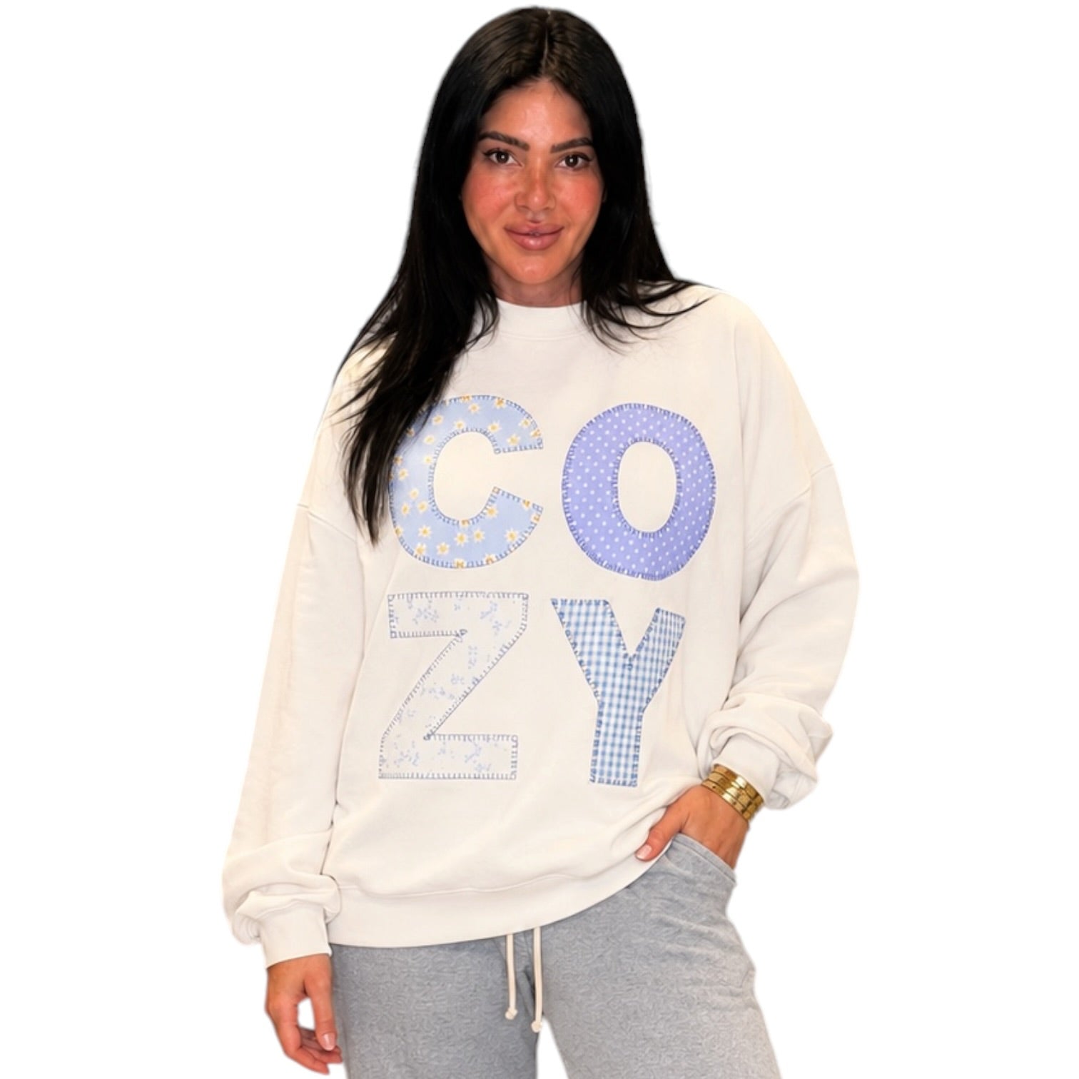 A woman modeling a cream-colored crewneck sweatshirt with 'COZY' written across the front in blue patterned letters, paired with gray sweatpants against a plain white background.