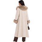 A woman shown from the back wearing a long cream-colored coat with a large brown and beige faux-fur collar and matching faux-fur trimmed cuffs, paired with dark brown pants and holding a small brown handbag against a plain white background.
