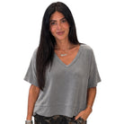 A woman with long black hair wearing a gray short-sleeve V-neck top and camouflage pants, standing against a plain white background.
