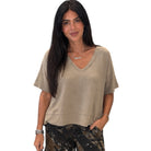 A woman modeling a beige short-sleeve V-neck top with a relaxed fit, paired with camouflage pants, against a plain white background.
