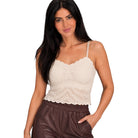 A woman modeling a cream-colored lace camisole top with thin straps and a scalloped neckline, paired with brown pants, against a plain white background.