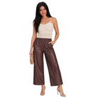 A woman modeling a cream lace camisole top and brown wide-leg faux leather pants, holding a small beige clutch against a plain white background.