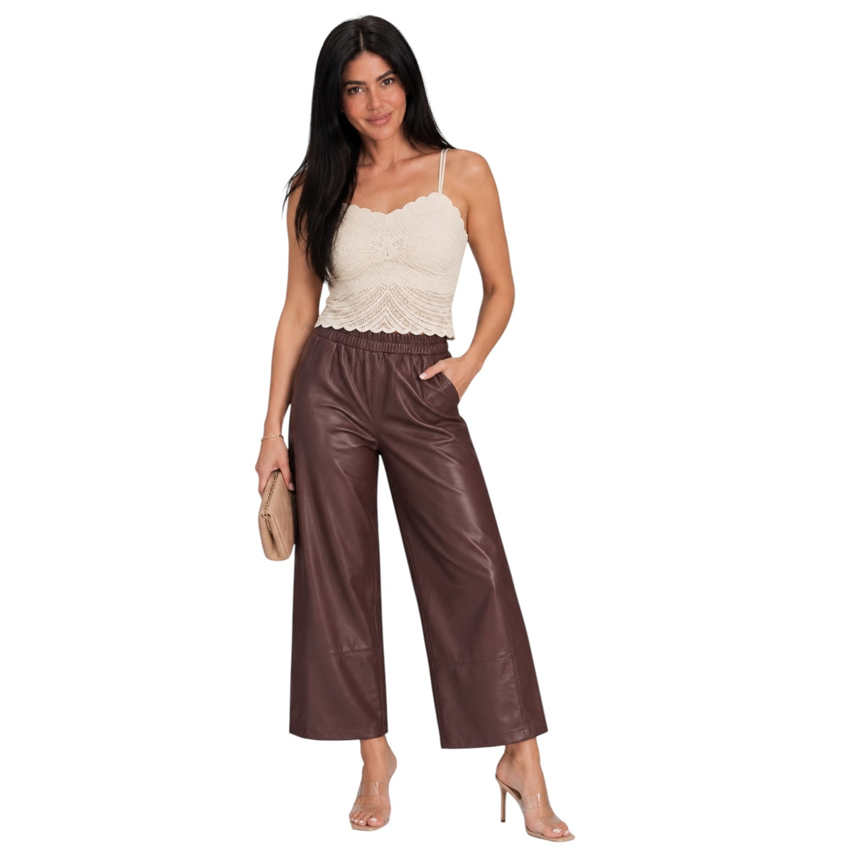 A woman modeling a cream lace camisole top and brown wide-leg faux leather pants, holding a small beige clutch against a plain white background.