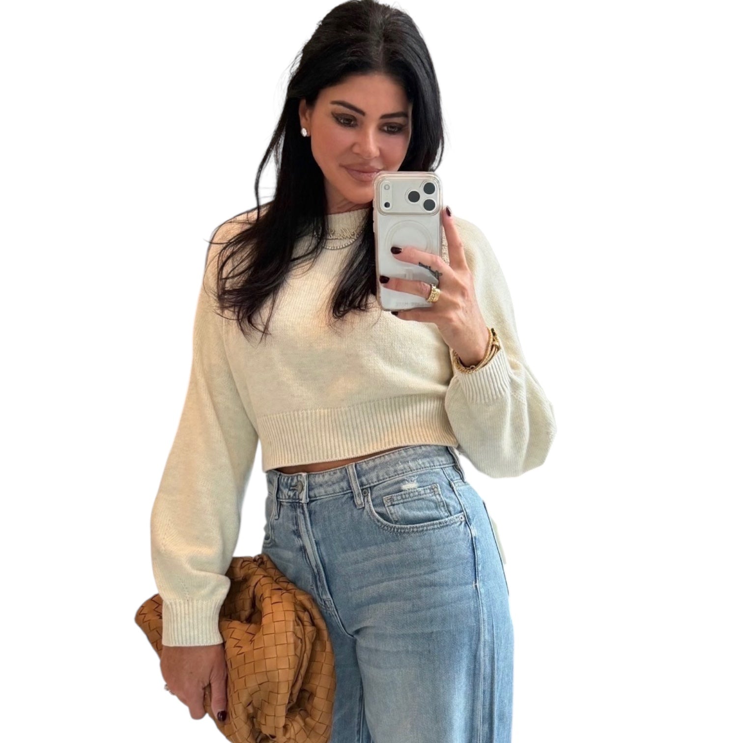 Woman taking a mirror selfie wearing a beige sweater and blue jeans on a white background