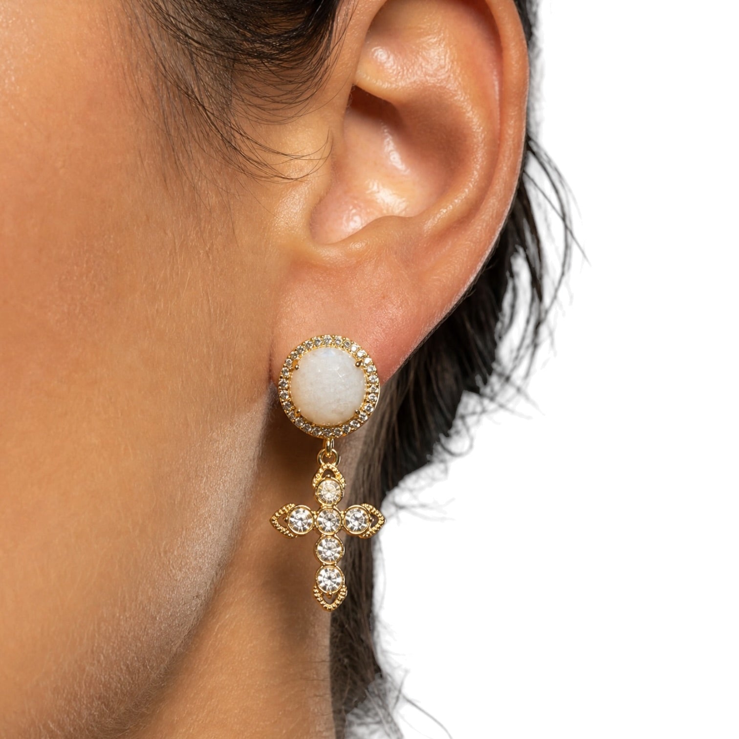 A close-up of a person's ear wearing a gold drop earring with a round white stone at the top and a dangling cross motif set with small clear stones.
