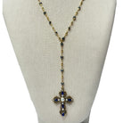 A gold-tone cross pendant necklace with blue gemstones and a central white pearl, displayed on a beige fabric jewelry bust against a white background.