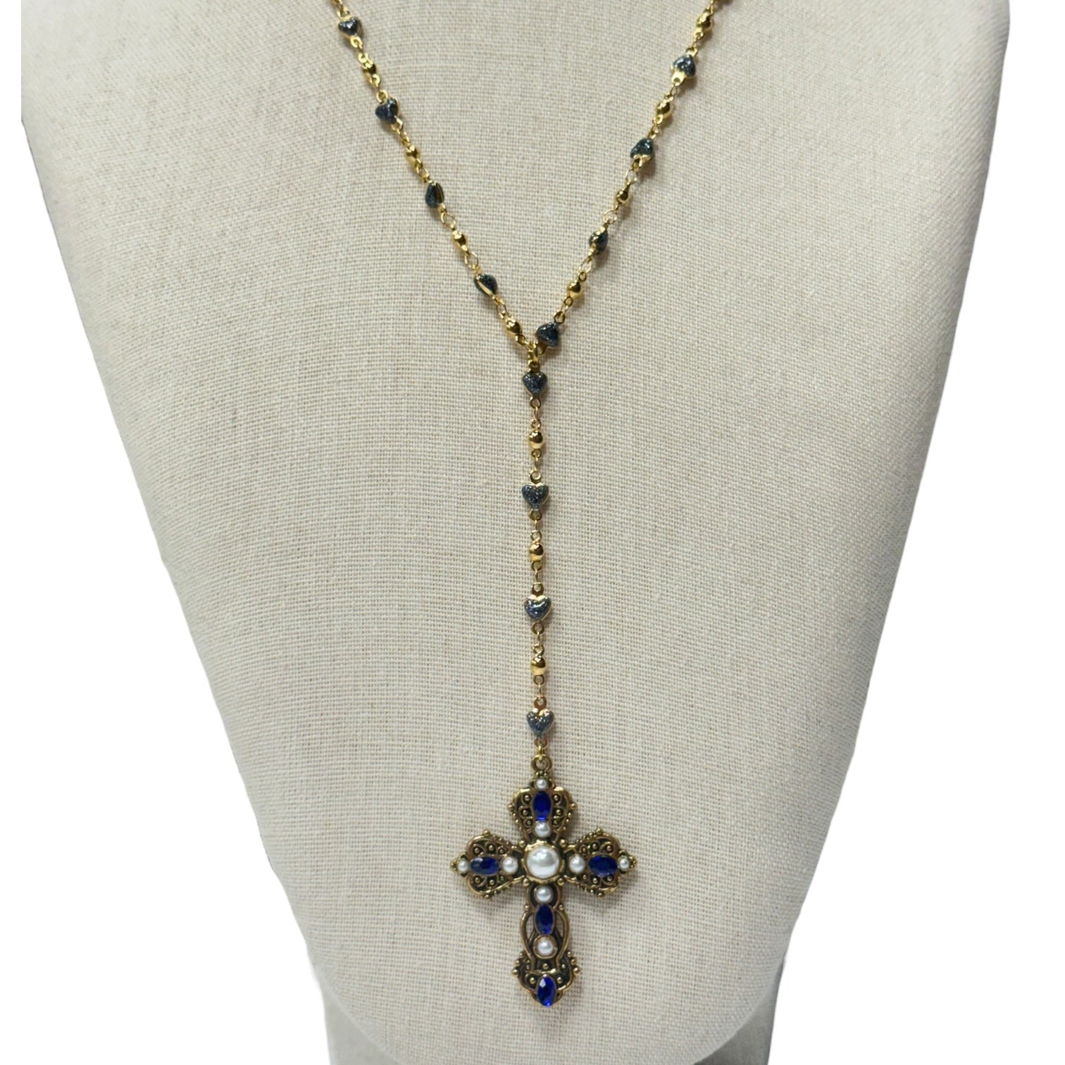 A gold-tone cross pendant necklace with blue gemstones and a central white pearl, displayed on a beige fabric jewelry bust against a white background.