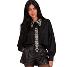 A woman modeling a black long-sleeve blouse with ornate silver beaded embroidery on the collar and front placket, paired with black shorts, against a plain white background.