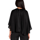 A woman viewed from the back wearing a black draped cape-style top with wide sheer sleeves and black pants, against a plain white studio background.