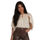 A woman modeling a cream-colored blouse with flutter sleeves and high-waisted, wide-leg brown faux-leather pants, holding a black woven handbag against a plain white background.