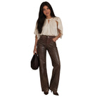 A woman modeling a cream-colored blouse with flutter sleeves and high-waisted, wide-leg brown faux-leather pants, holding a black woven handbag against a plain white background.