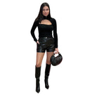 Woman wearing a black sweater with leather shorts and boots, holding a matching handbag on a white background.
