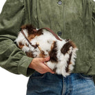A person wearing an olive green corduroy jacket and blue jeans holds a brown and white fur shoulder bag with a brown leather strap.