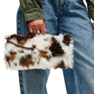 A person wearing blue jeans and an olive green jacket holds a fluffy faux-fur clutch bag with brown and white cow-print patches and a brown leather strap with gold hardware.