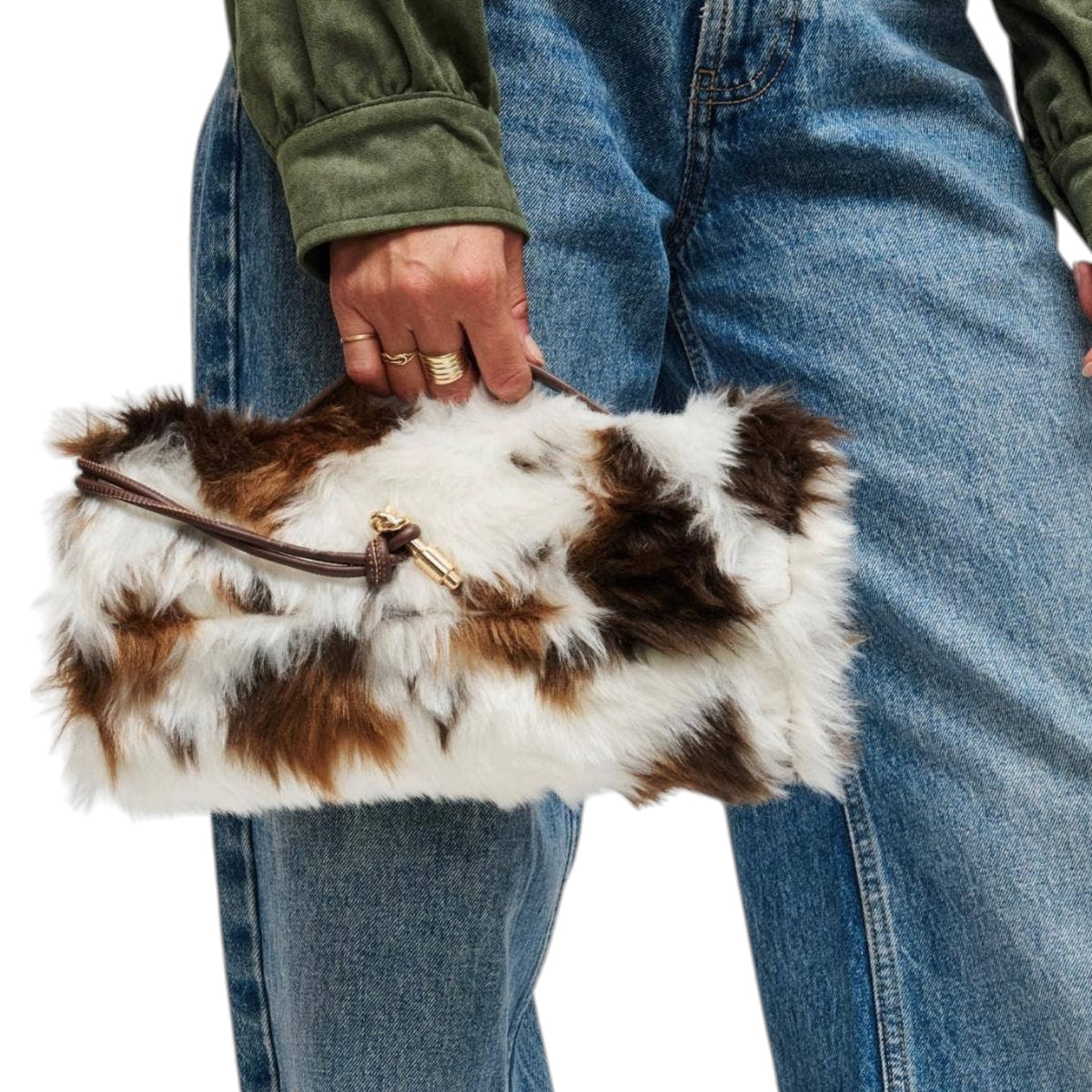 A person wearing blue jeans and an olive green jacket holds a fluffy faux-fur clutch bag with brown and white cow-print patches and a brown leather strap with gold hardware.
