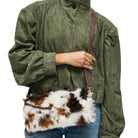 A person wearing an olive green textured jacket and blue jeans carries a brown and white cow-print faux fur shoulder bag with a brown strap.