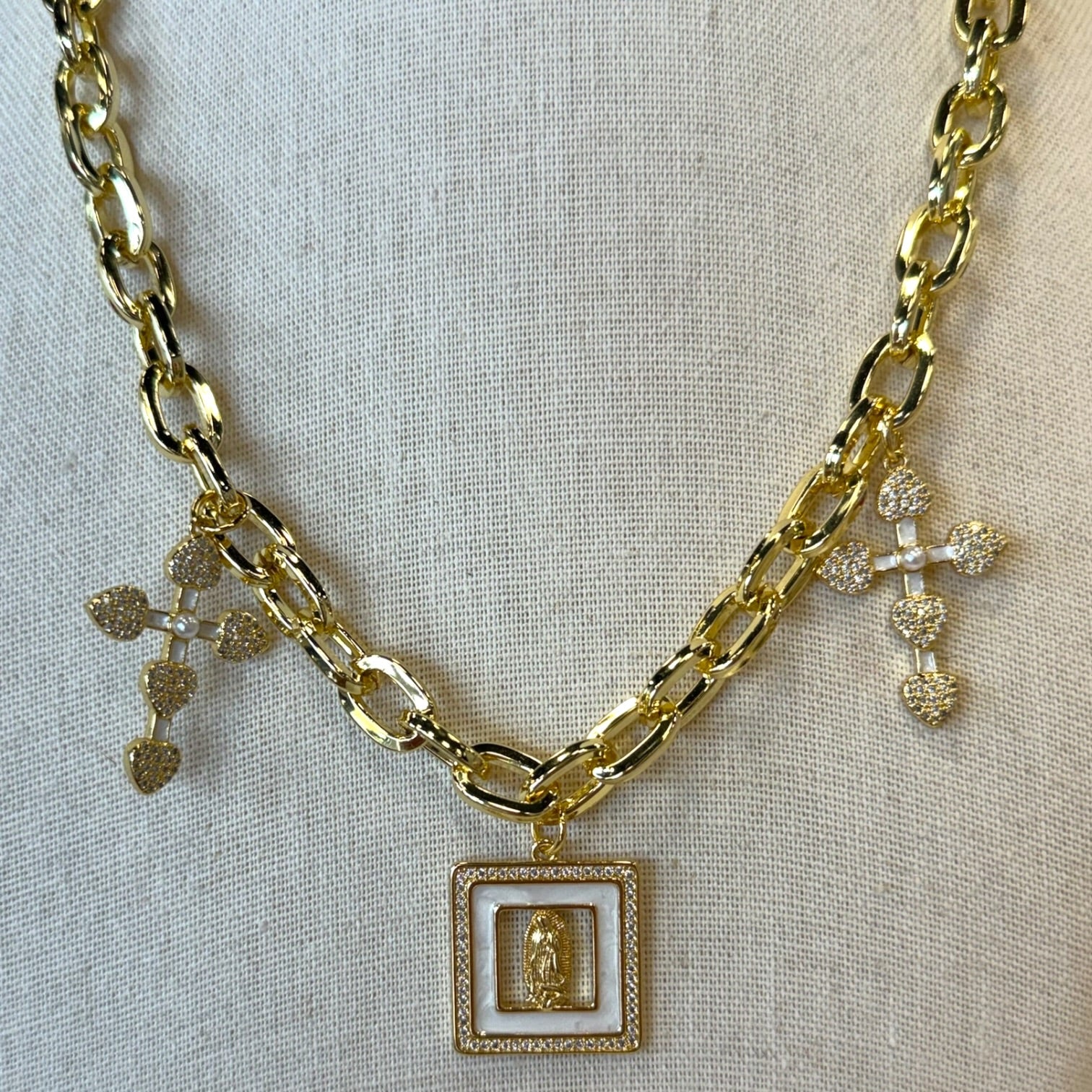 A gold-toned multi-strand chain necklace displayed on a textured beige fabric background. The necklace features three distinct pendant styles: a textured gold leaf charm, a gold cross encrusted with clear rhinestones, and a square gold pendant with a white inlay and a small gold figure in the center.