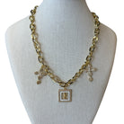 A gold-tone chain necklace displayed on a cream fabric jewelry bust. The necklace features a central square pendant with a gold cross and a white inlay, flanked by two small gold cross charms with clear rhinestone details.