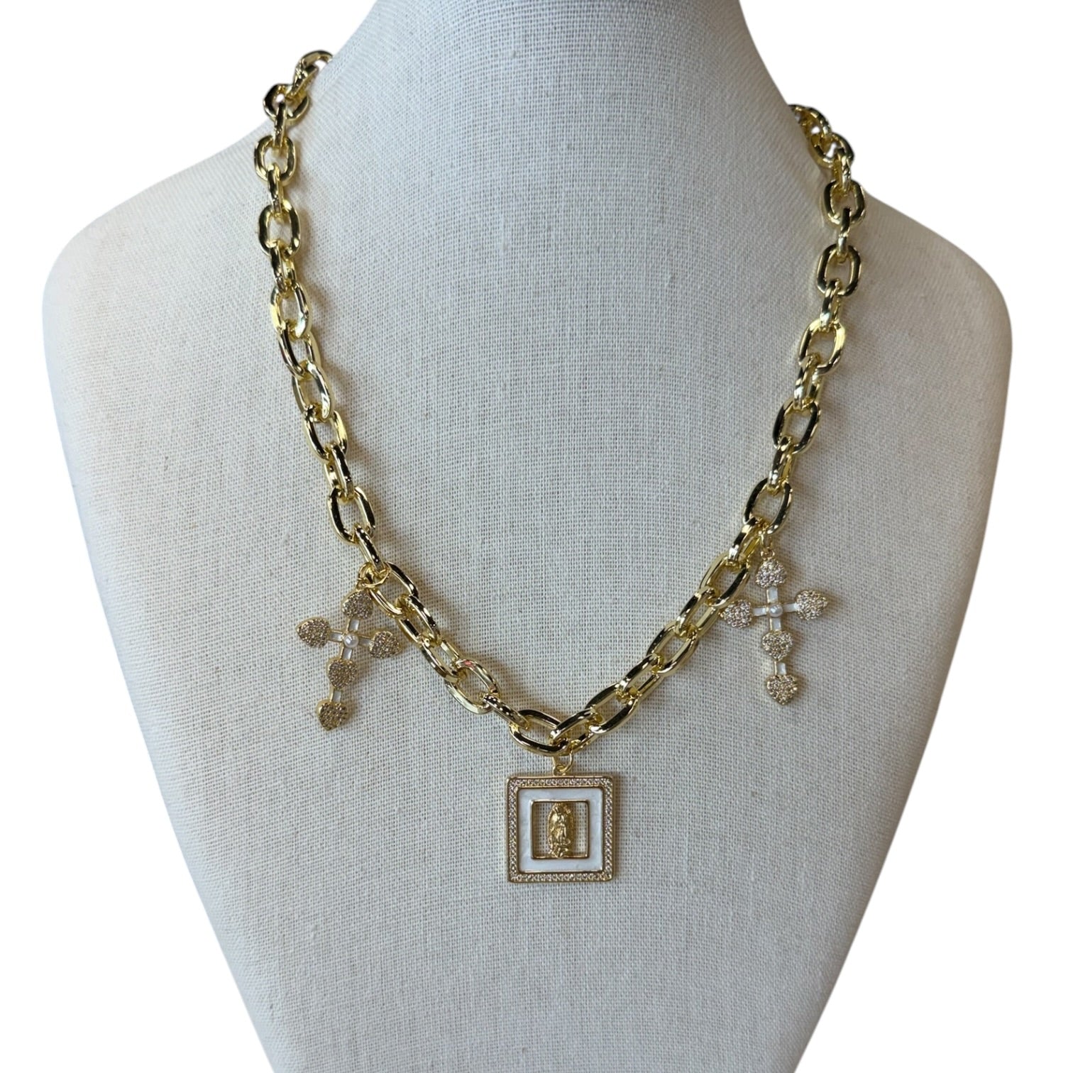 A gold-tone chain necklace displayed on a cream fabric jewelry bust. The necklace features a central square pendant with a gold cross and a white inlay, flanked by two small gold cross charms with clear rhinestone details.
