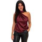 A woman modeling a satin-like one-shoulder top in deep burgundy, paired with black faux-leather pants, against a plain white background.