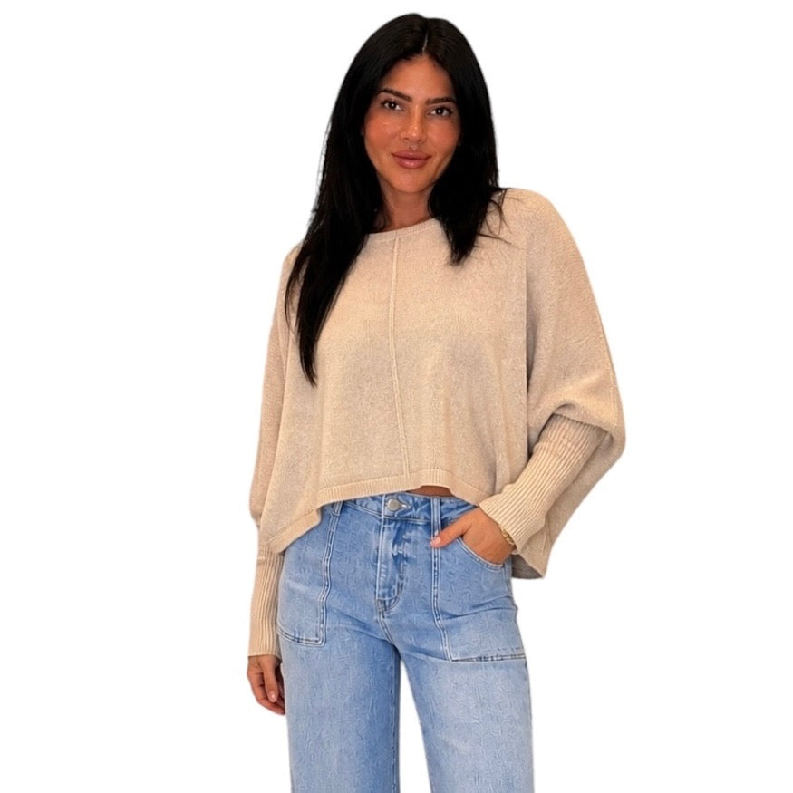 A woman modeling a light beige knit sweater with long draped sleeves and a relaxed fit, paired with light blue jeans, against a plain white background.