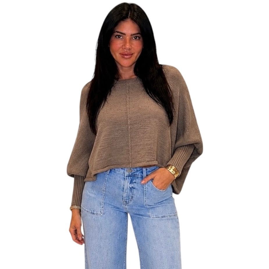 A woman modeling a taupe brown knit sweater with balloon sleeves and light blue jeans against a plain white background.