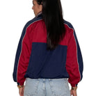 A woman shown from the back wearing a colorblock windbreaker jacket with navy blue and red panels, light blue jeans, and a black ponytail against a white background.