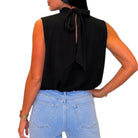 A woman shown from the back wearing a sleeveless black blouse with a large tie-back detail and light blue jeans, against a plain white background.