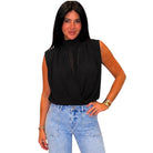 A woman modeling a sleeveless black blouse with a high mock neck and twisted front detail, paired with light blue jeans, against a plain white background.