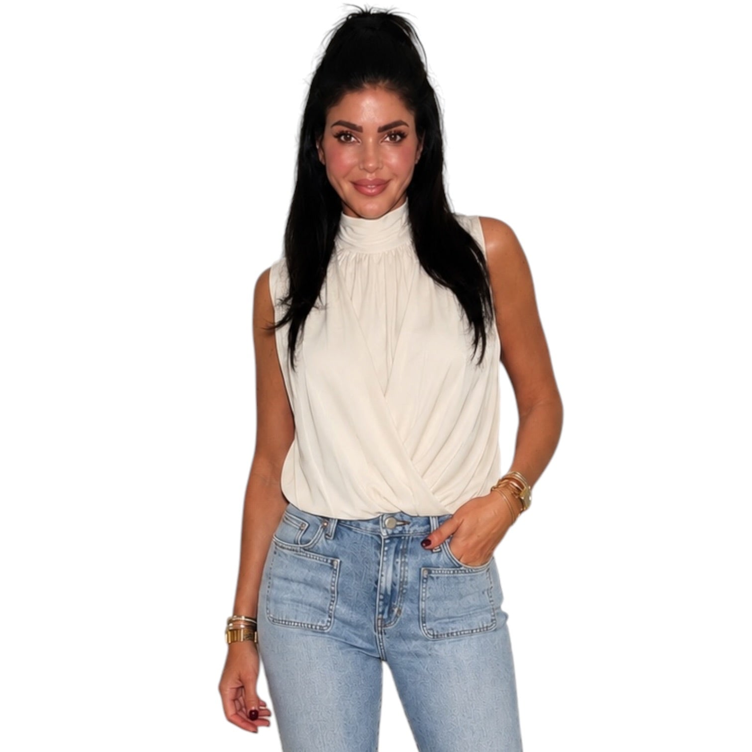 A woman modeling a sleeveless cream-colored mock neck top with a twisted front detail, paired with light-wash blue jeans, against a plain white background.