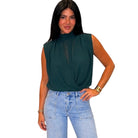 A woman modeling a sleeveless dark green mock-neck blouse with a sheer mesh panel at the chest, paired with light-wash blue jeans, against a plain white background.