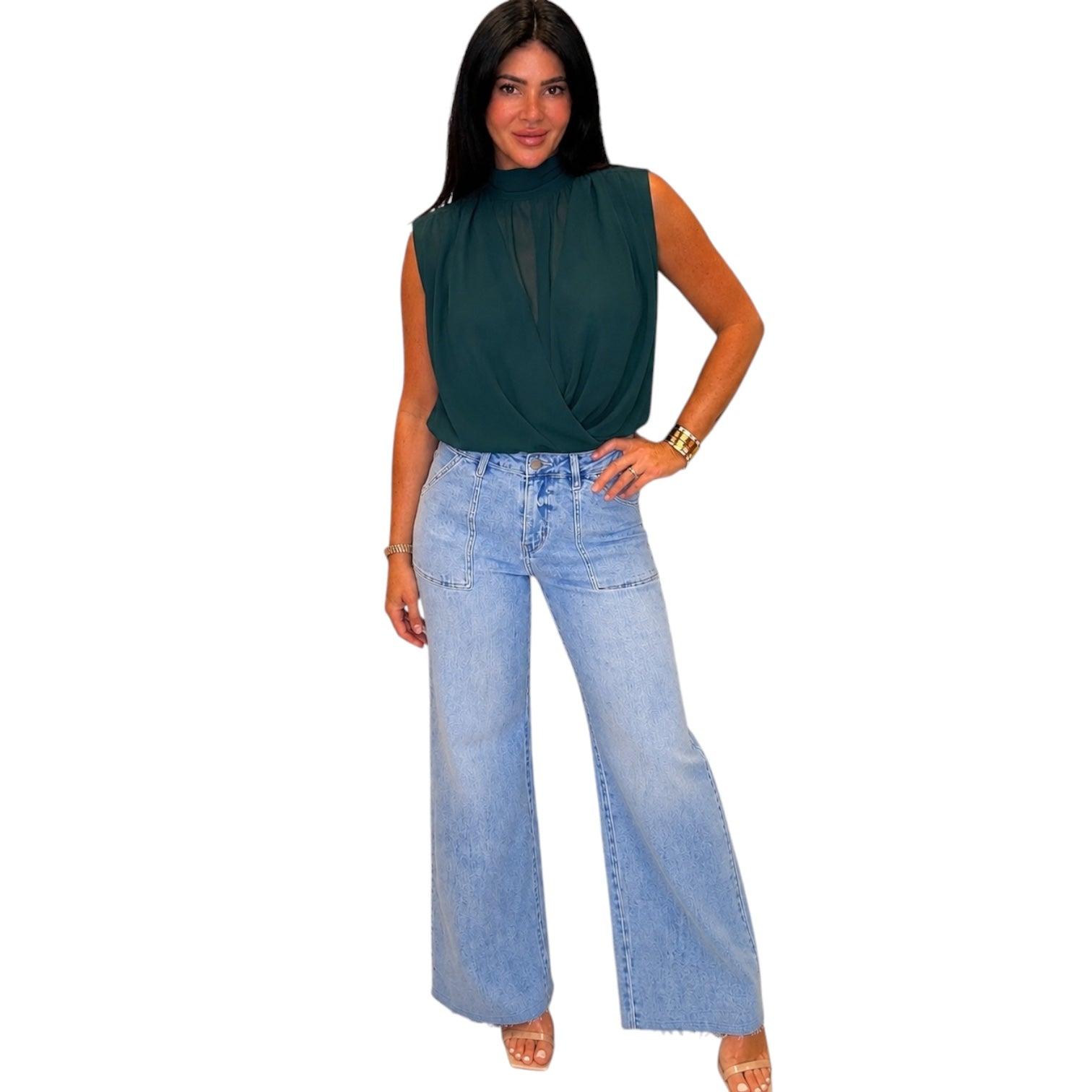 A woman modeling a sleeveless dark green top with a high neck and twisted front detail, paired with light-wash wide-leg denim jeans, against a plain white background.