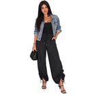 A woman modeling a black wide-leg jumpsuit with a drawstring waist and side tie details, styled with a light blue denim jacket, black tank top, and black strappy heels against a plain white background.