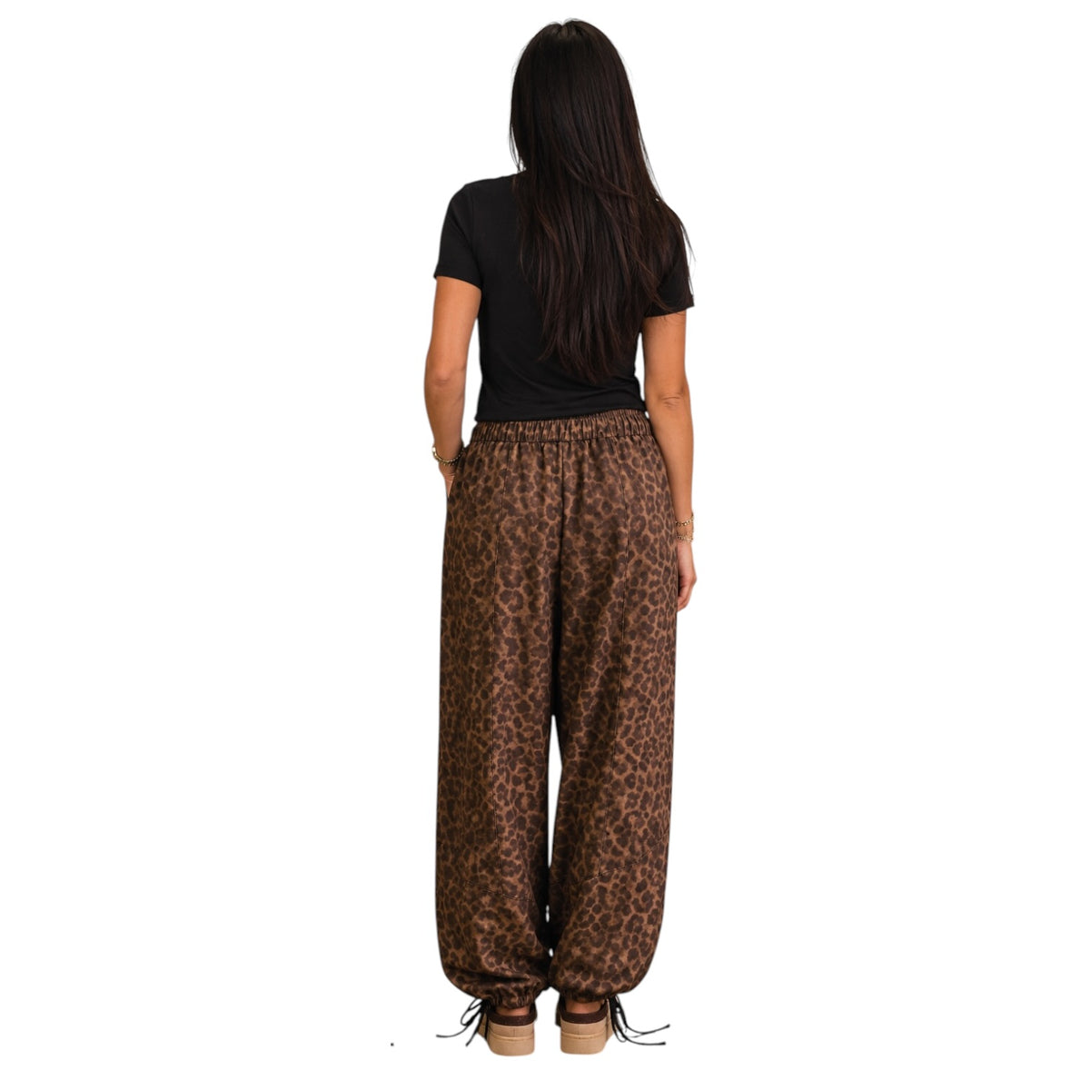 A woman viewed from behind wearing loose brown leopard-print pants, a black short-sleeve top, and brown platform sandals against a plain white background.