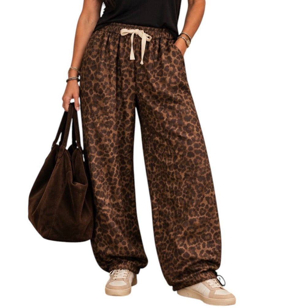 A woman modeling loose-fit leopard-print pants with a cream drawstring, paired with a black V-neck T-shirt and beige sneakers, holding a brown tote bag against a plain white background.