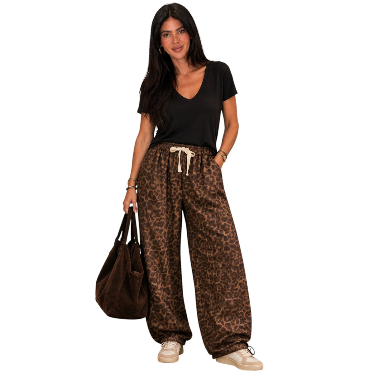 A woman modeling loose-fit leopard-print pants with a cream drawstring, paired with a black V-neck T-shirt and beige sneakers, holding a brown tote bag against a plain white background.