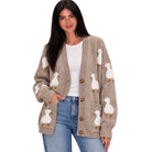 A woman wearing a beige knit cardigan with white duck motifs and brown buttons, styled over a white top and blue jeans against a plain white background.
