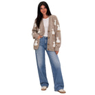 A woman modeling a beige cardigan with white goose motifs, paired with a white t-shirt, wide-leg blue jeans, and white sneakers, set against a plain white background.
