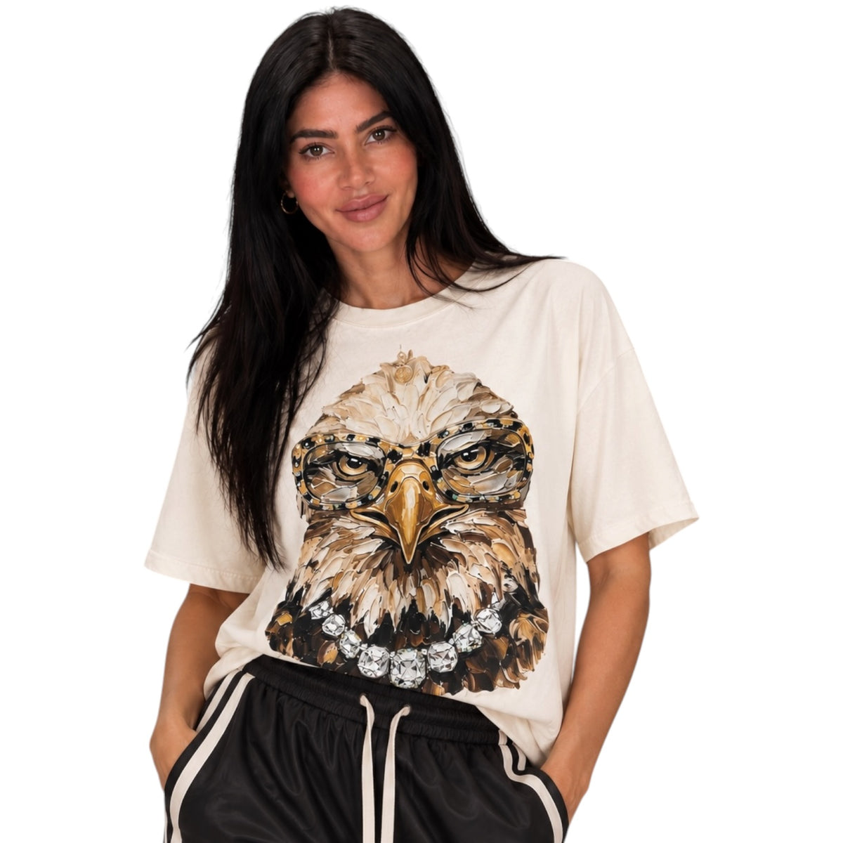 A woman modeling a cream-colored oversized t-shirt with a detailed graphic of a bald eagle wearing a diamond necklace, paired with black athletic shorts against a solid white background.