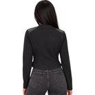 A woman viewed from the back wearing a black cropped long-sleeve jacket with dark gray shoulder panels and dark gray jeans, set against a plain white background.