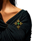 A close-up of a woman wearing a black draped top and wearing a gold brooch with a green oval center stone on her shoulder.